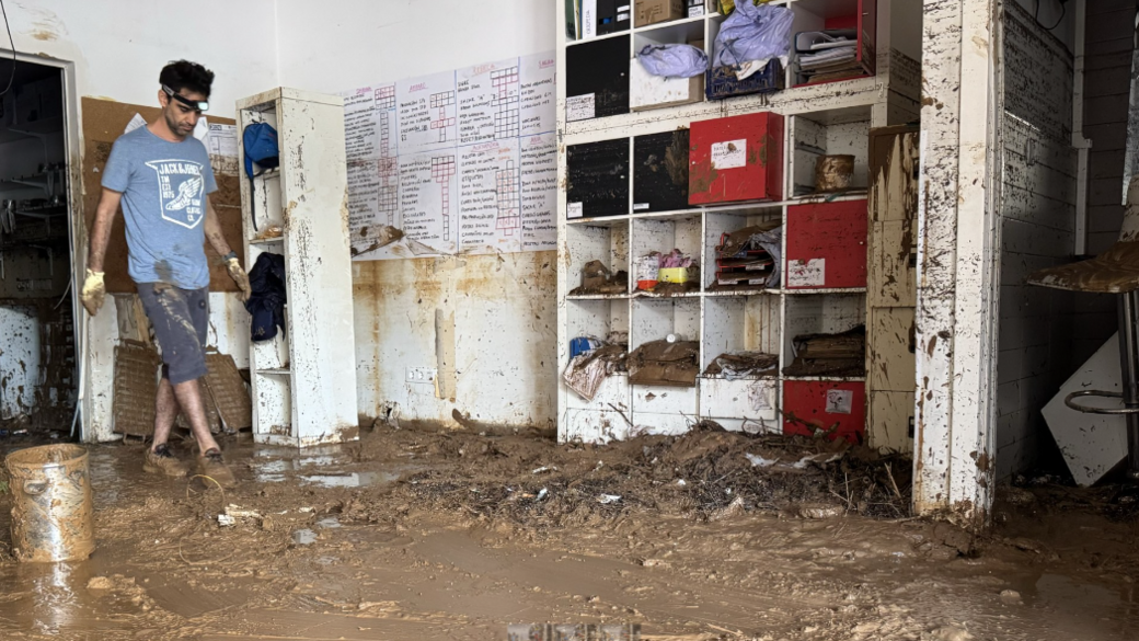 Pharmacist Miguel wearing mud-caked gloves and shoes and a head torch steps through his mud-drenched shop after the floods