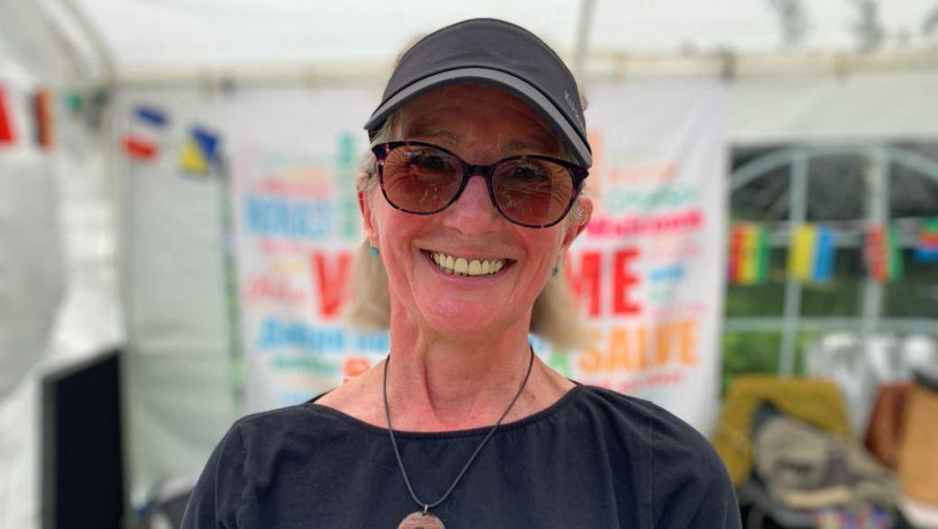 Stephanie Gray is wearing sunglasses and a black cap, smiling wearing a black top - standing in front of a marquee with colourful bunting and signage.