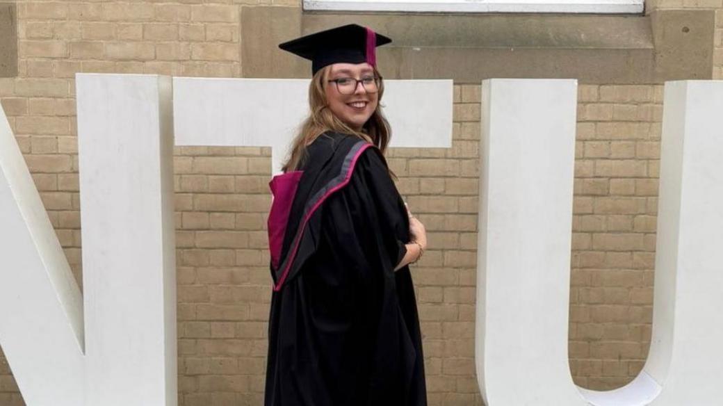 Stephanie Irons wearing a graduation gown and cap in front of a sign for Nottingham Trent University