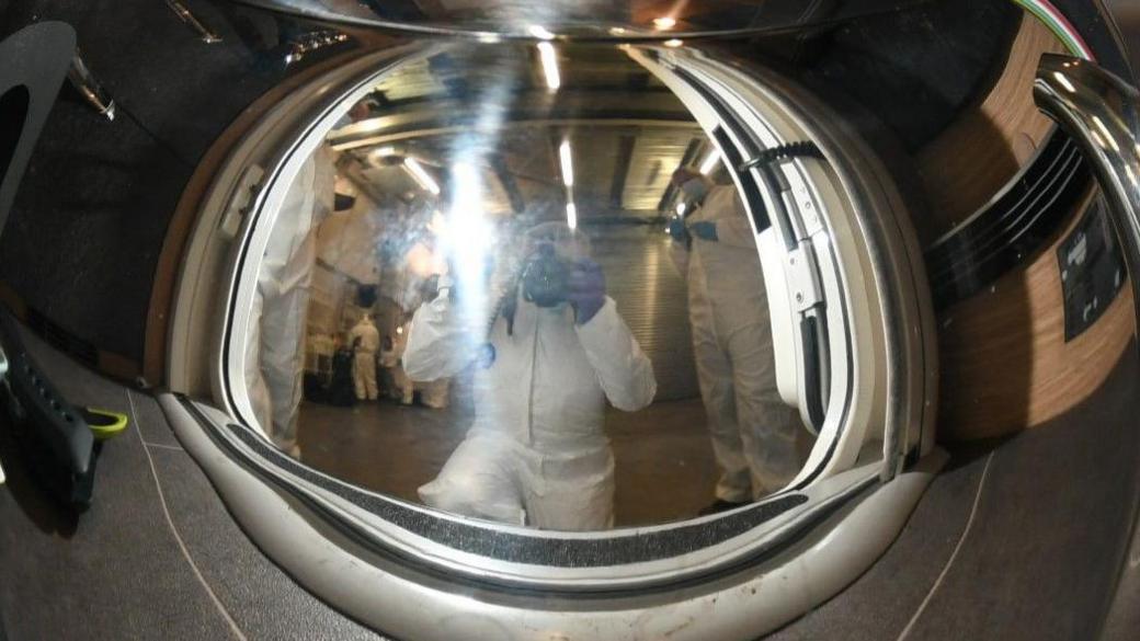 A stainless steel teapot. The reflection shows a man taking a photograph of the teapot. 