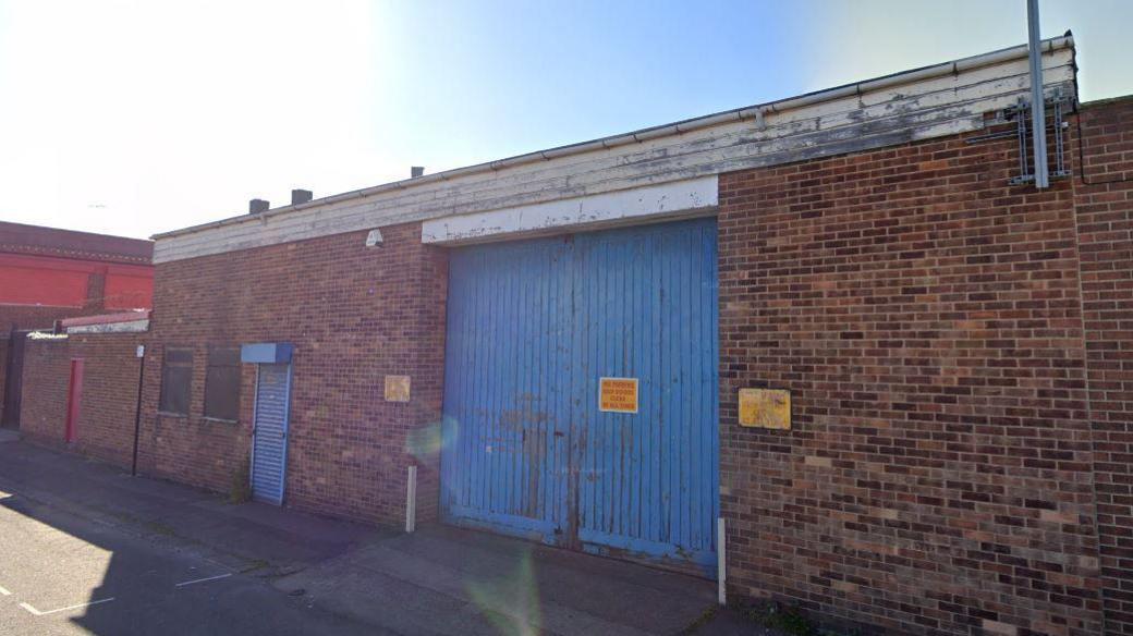 A wooden blue double door, part of a brick, single-storey building. It has a small ramp for entry from the street.