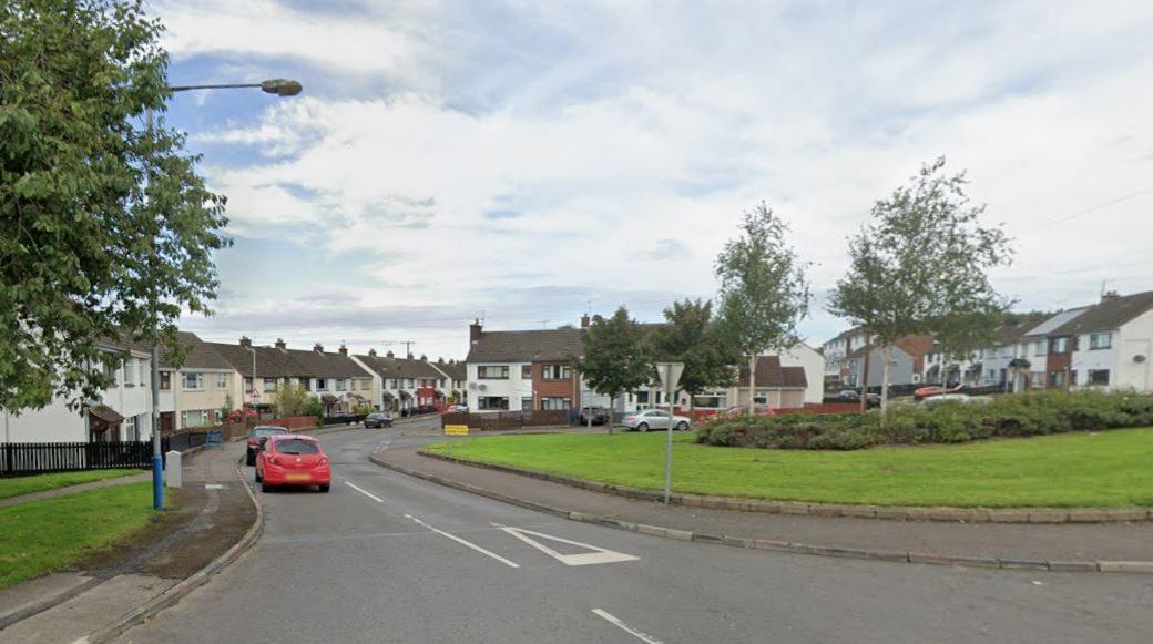 An image of an entrance to a street. Near the entrance are a number of terraced houses, as well as a green area made up of grass, bushes and some trees. Parked cars can been along the street