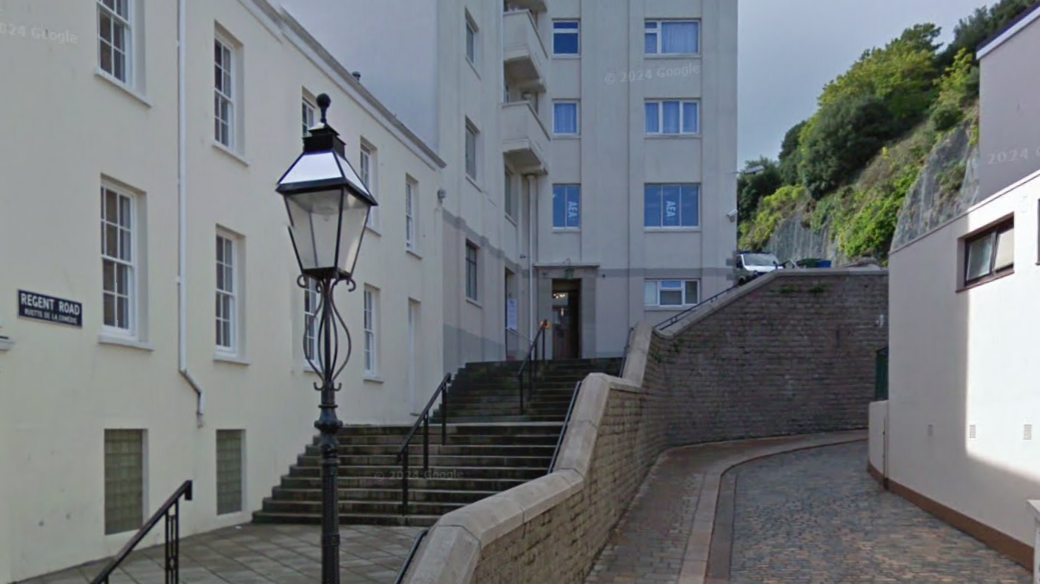 A set of stone steps leading up to a door for the Regent House. There is a sign on the wall that reads REGENT ROAD and underneath RUETTE DE LA COMEDIE. To the right of the steps is a wall and a path next to a building. There is an ornate iron work lamp post in the foreground.