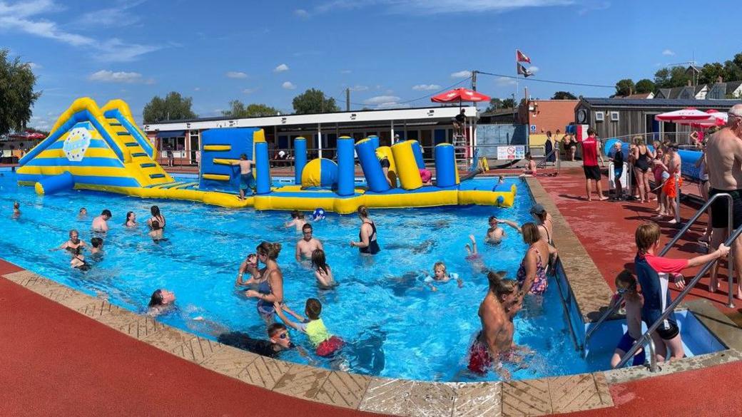 Beccles Lido is a large open-air pool. Lots of people of all ages are enjoying the water. There is also an inflatable yellow and blue obstacle course to one side of the pool