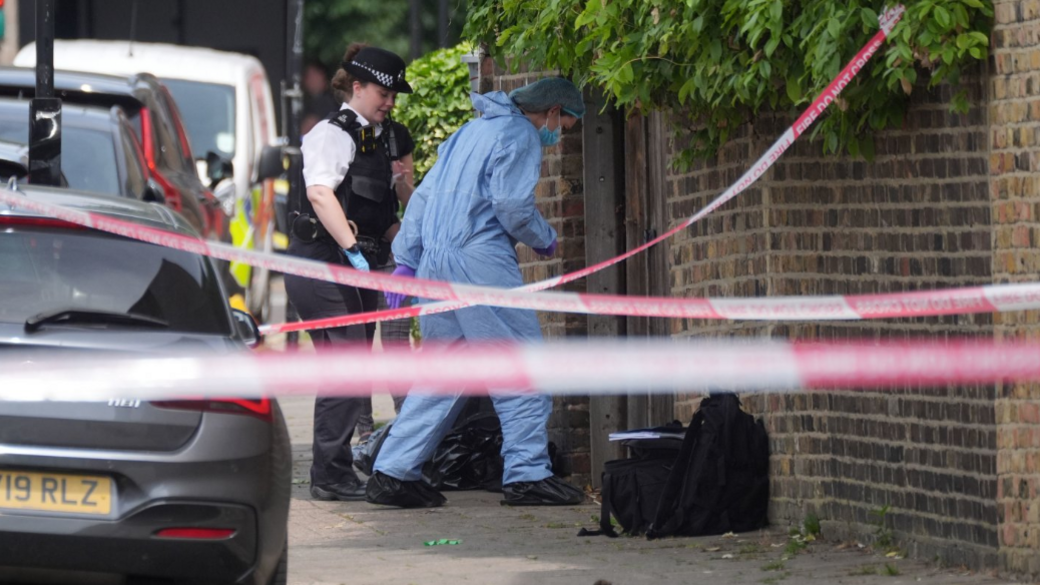 A forensic investigator in a blue suit and face mask examines a scene behind police tape, with a uniformed officer and police vehicles nearby.