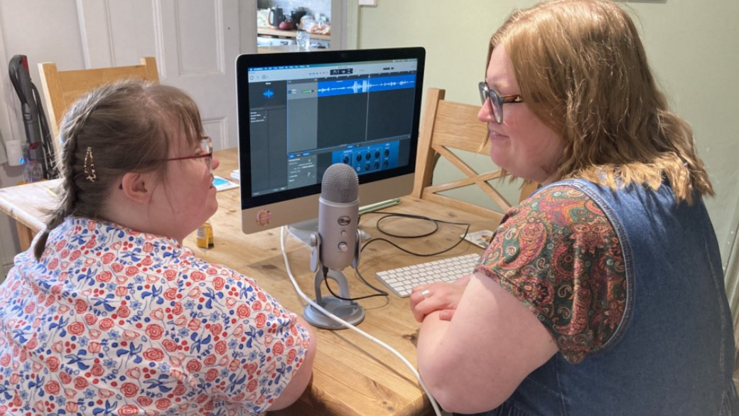 Lucy and Sarah sitting at a desk with a microphone and a computer on it