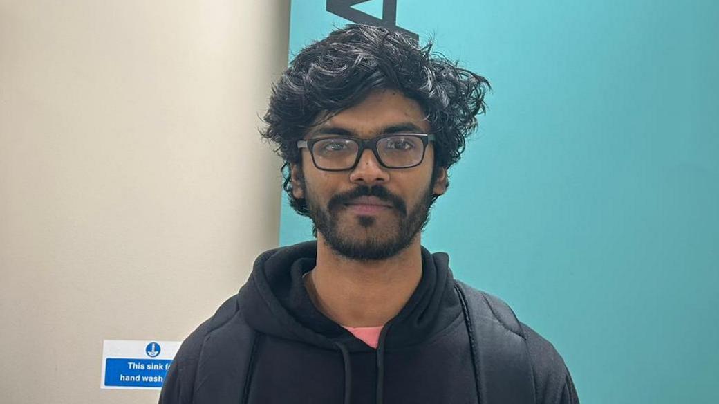 Muphuselban stands in front of a blue background. He has black hair and black glasses. He has a black moustache and beard. He is wearing an orange top with a black hoodie.