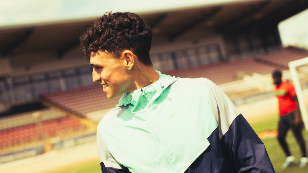 Phil Foden, in a largely light green zip-up top, looks to his right with a smile as he attends Rico Lewis' charity football tournament