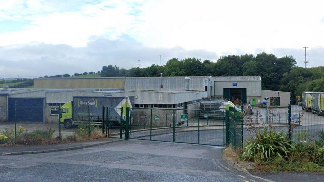 A grey factory with a collection of green and black lorry's and vans sitting in the forecourt, it is sorrounded by green metal fencing