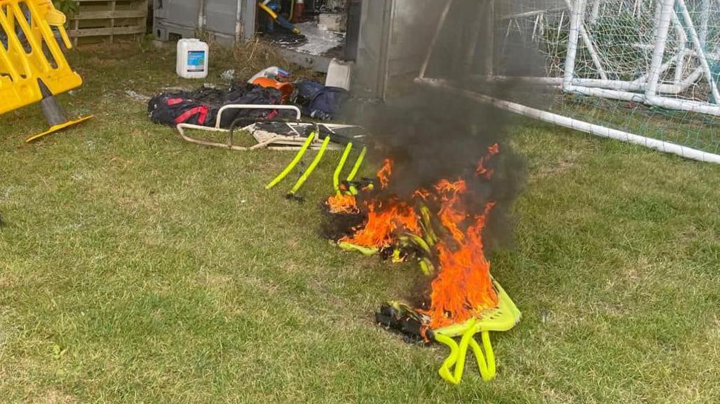 A yellow pop-up football goal on fire laying on the grass outside a storage container. There is other equipment strewn all over the grass and spilling out of the container. There is black smoke coming off the burning goal.