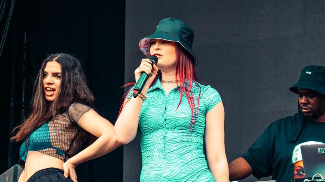 Three women and a man are performing on a festival stage. The woman in the centre has on a turquoise jumpsuit with wave patterns on and a bucket hat. She has long red hair and is singing into a microphone. The two women to her side are wearing bikinis to match her dress and black trousers. A man is mixing at decks in the background. 