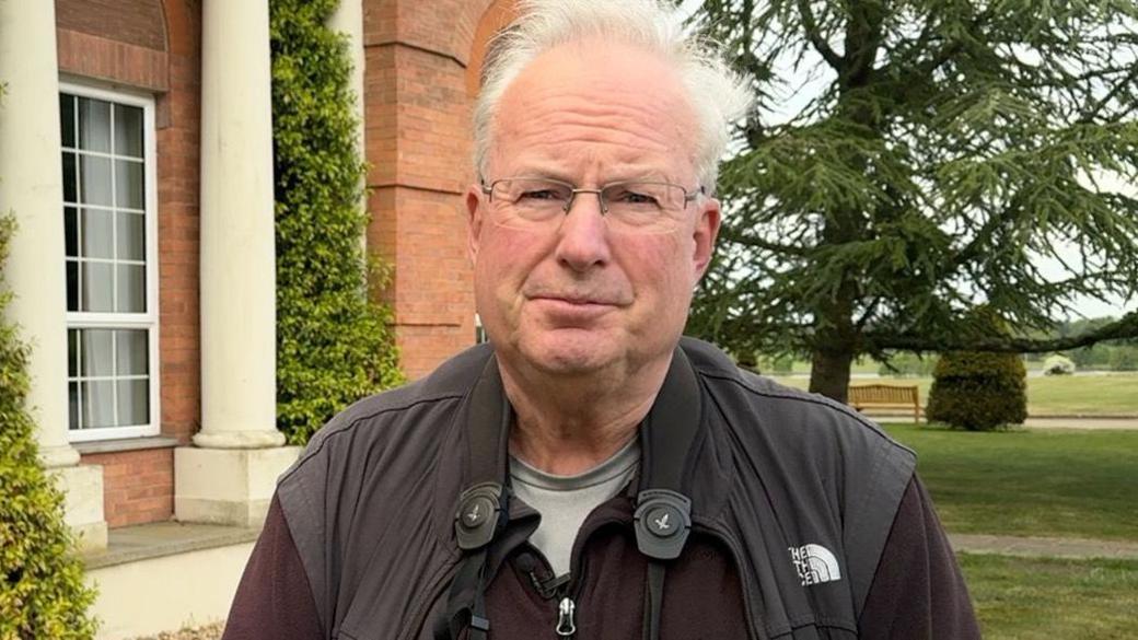 A man is standing looking at the camera wearing glasses. He has binoculars around his neck and is wearing a black coat. There is a red-brick building in the with white pillars and greenery in the background.