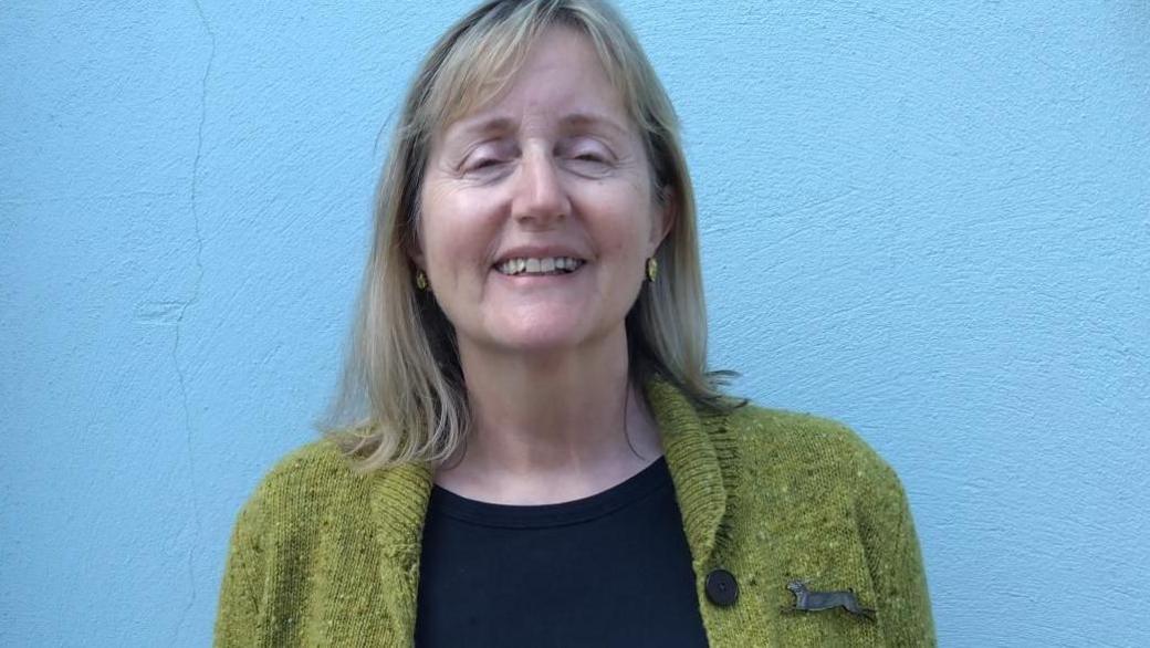 Heather Hurt, a woman with a friendly smile, she has a fringe and brown/grey hair, stood in front of a blue wall.