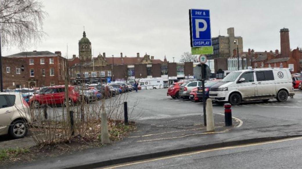 A city centre car park, filled with parked vehicles. A sign indicates a pay-and-display scheme is in operation.
