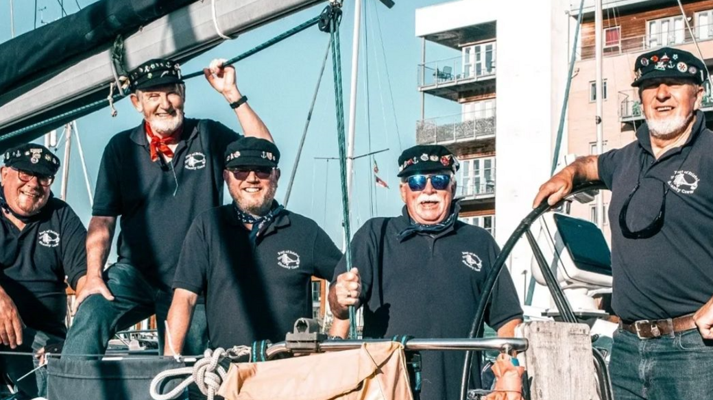 Five sailors all in blue polo shirts with a logo and peaked caps with badges on, smiling to camera from a sailing boat with blue skies and a block of flats behind them