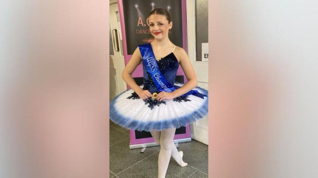 A photo of Eleanor Hague. She has brown hair in a bun and is wearing a tutu and is smiling