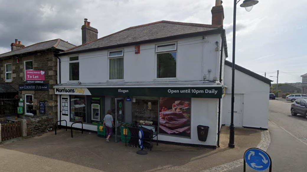 A Morrisons shop in a two-storey white painted building with a side entrance andlean-tp next to a road at the end of a line of shops.