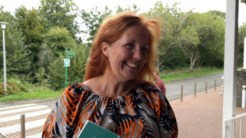 A smiling, red-haired woman win a patterned dress stands in front of a road and a zebra crossing.