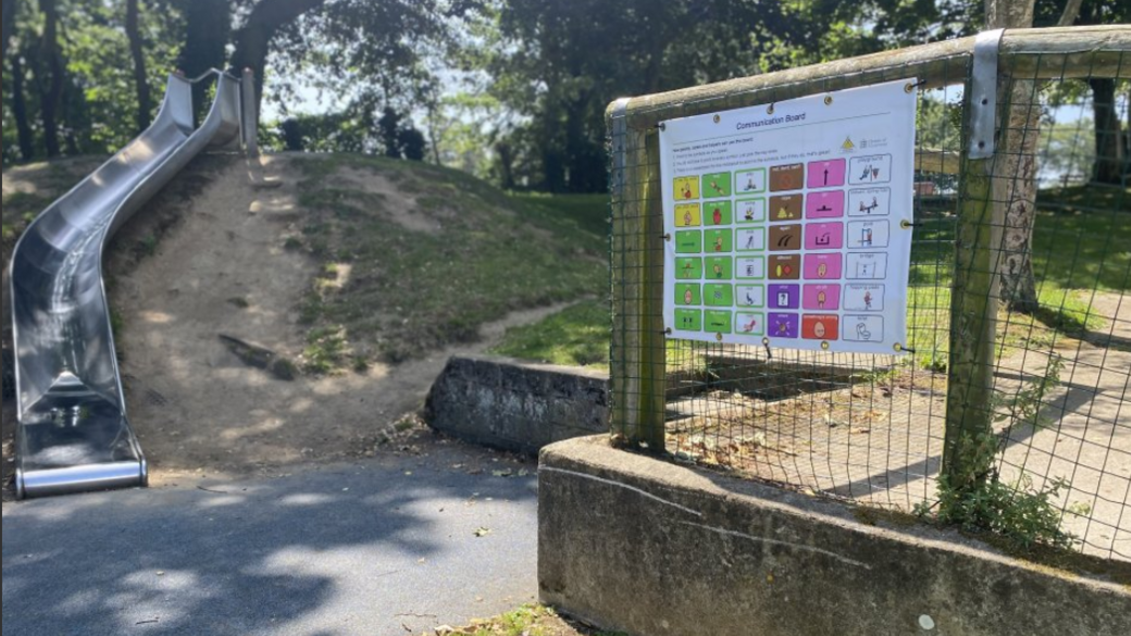 A large communication poster on a fence on the right at a play park. There is a slide on the left which is on a grass bank.