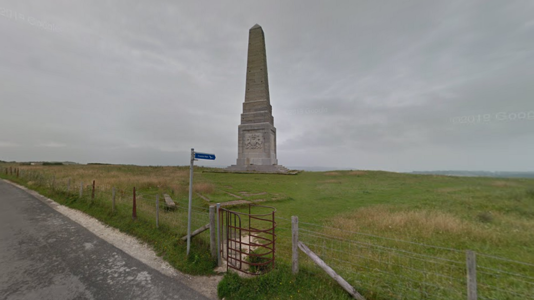 A stone monument stands in the middle of a grassy field. It is enclosed by a wire fence, which has an iron gate. The sky is grey.