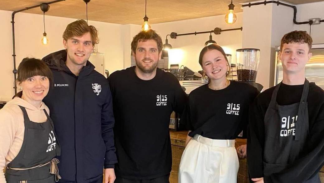 Five people in branded t-shirts and aprons stand together smiling in a café.