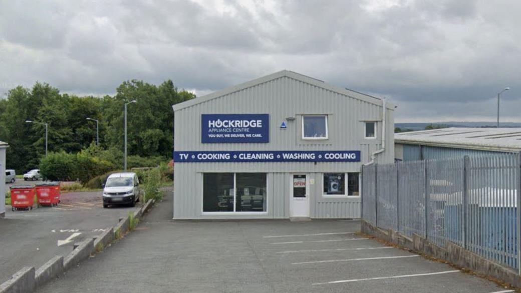A grey corrugated-iron building in an industrial estate. A sign reads Hockridge Appliance Centre while another reads: TV, Cooking, Cleaning, Washing, Cooling.