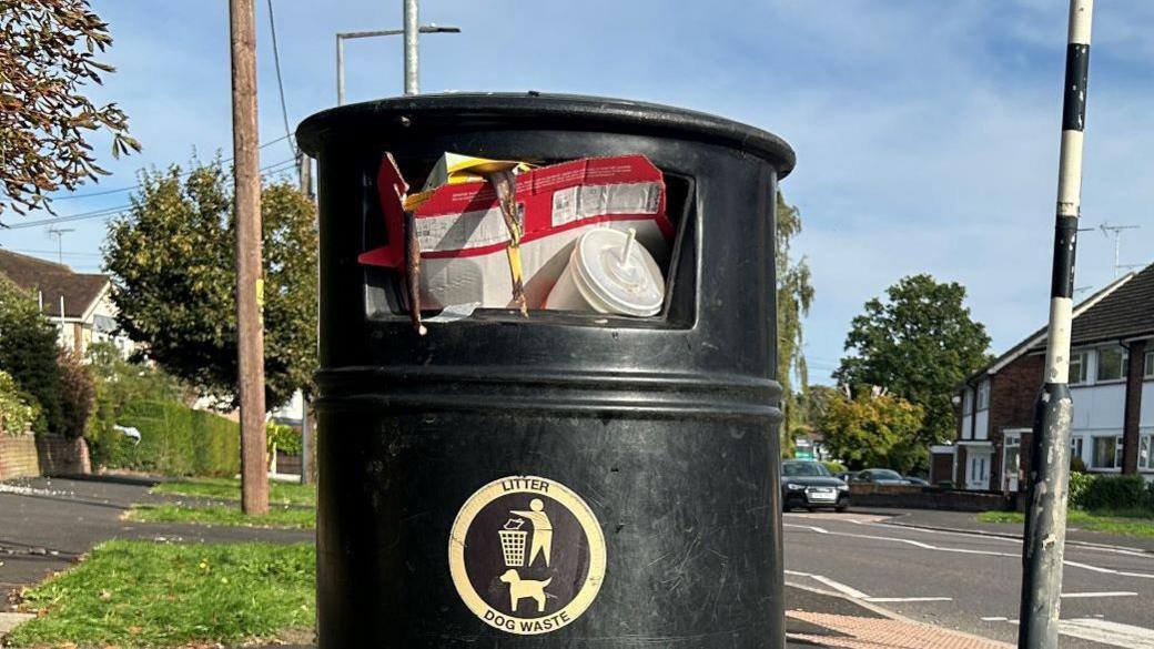 A black bin in Brentwood that has cardboard and a takeaway drink cup among the rubbish close to falling out. The bin is next to a zebra crossing on a residential road.