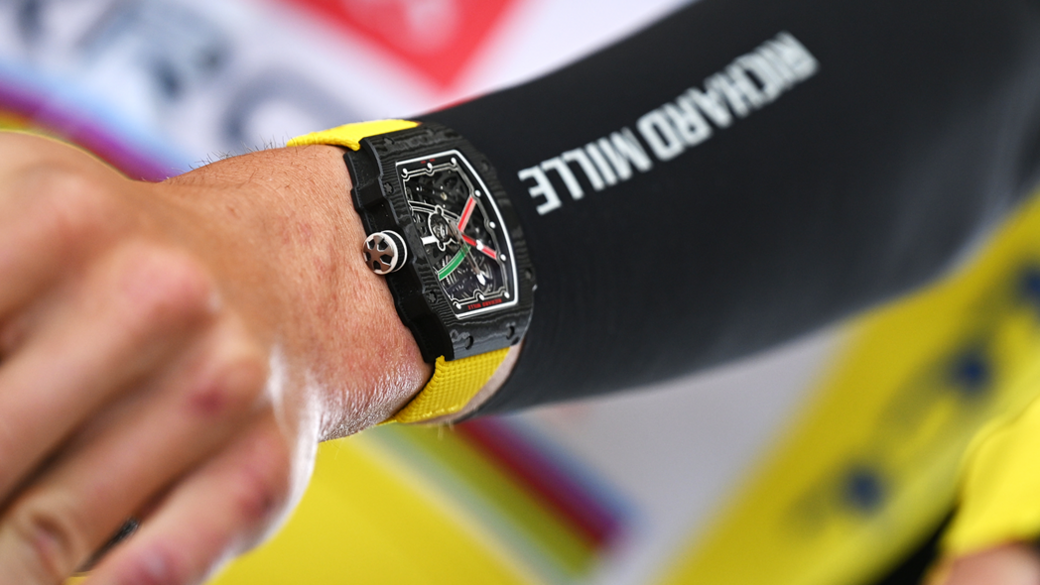 A close-up of a square watch with a black face and a yellow strap on the arm of a cyclist, who wears a Richard Mille sponsorship logo on his sleeve.