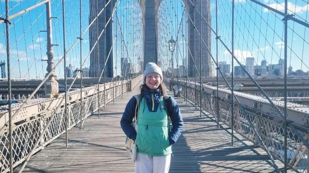 Becky Burke in a blue woolly hat and green jacket standing on the Brooklyn Bridge in New York