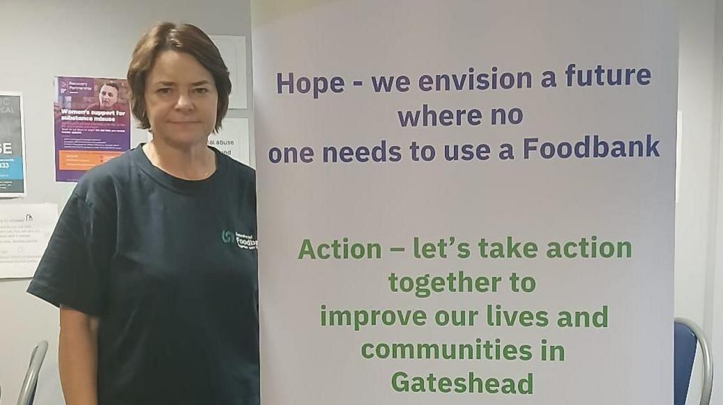 Diane Kindred standing next to a Gateshead Foodbank banner. She has dark hair and is wearing a dark blue T-shirt. The banner includes a number of action points such as "Hope - we envision a future where no one needs to use a foodbank".