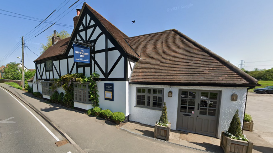 A black and white timber-framed pub that sits next to a road. In the background is a car park. A blue sign reads "The Navigation Inn"