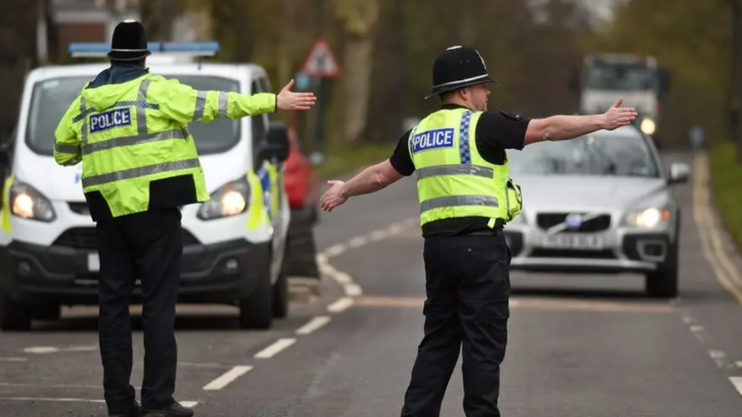 Two policemen in the foreground with their backs to camera, wearing hi-vis jackets and helmets, standing on a road and directing traffic with their arms. Their police van is visible on the left. A silver car is approaching them and a lorry is behind, quite blurred.