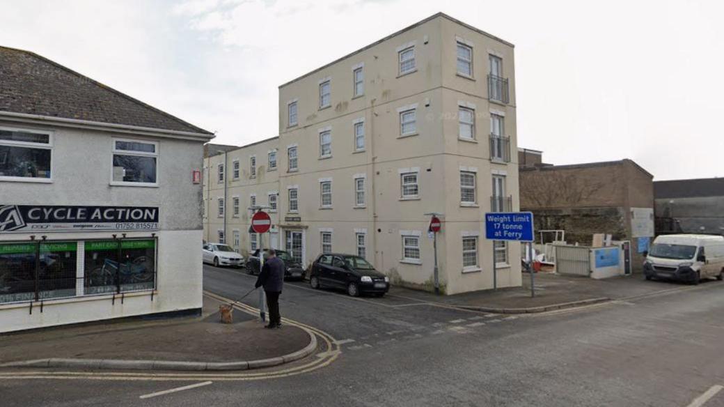 A Google Street View of the junction of Quarry Street and Harvey Street in Torpoint. A man is stood on the pavement looking down Quarry Street on the left of the image while walking a dog. A bike shop called Cycle Action is on the corner of the junction. A cream-coloured building is on the other side of the road. A blue sign saying "Weight limit 17 tonne at Ferry" is on the road. Several cars and a white van are parked on the roads.