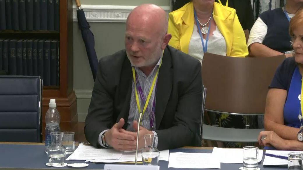 Chris Hagan is bald on top with grey hair at the sides and a grey beard. He is wearing a suit jacket and open-necked shirt with a yellow lanyard around his neck