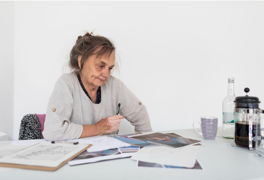 Artist Kaarina Kaikkonen is sat down looking at plans for her work on the table in front of her.