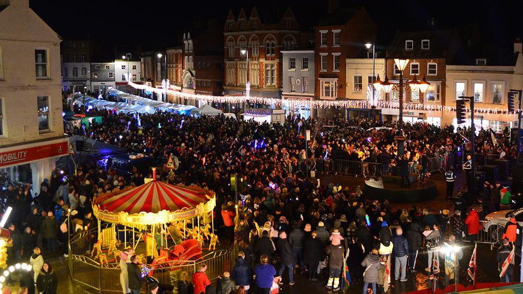 A night-time scene of crowds of people in a market square with a lit-up carousel ride in the corner