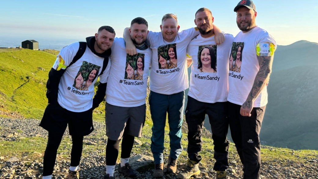 Mr Broadhead standing with his arms around friends on Snowdon. They are all wearing white T-shirts with his wife's picture on 