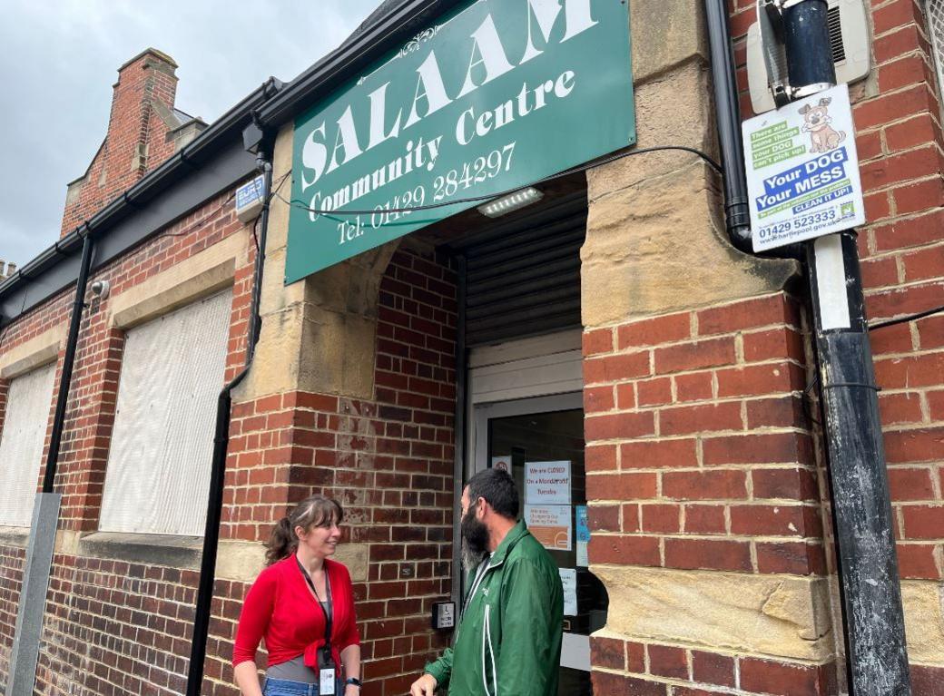 A woman with dark hair and a red top and a man with a green jacket stand talking to each other. They are outside a brick building with boarded up windows and a big sign saying Salaam Community Centre