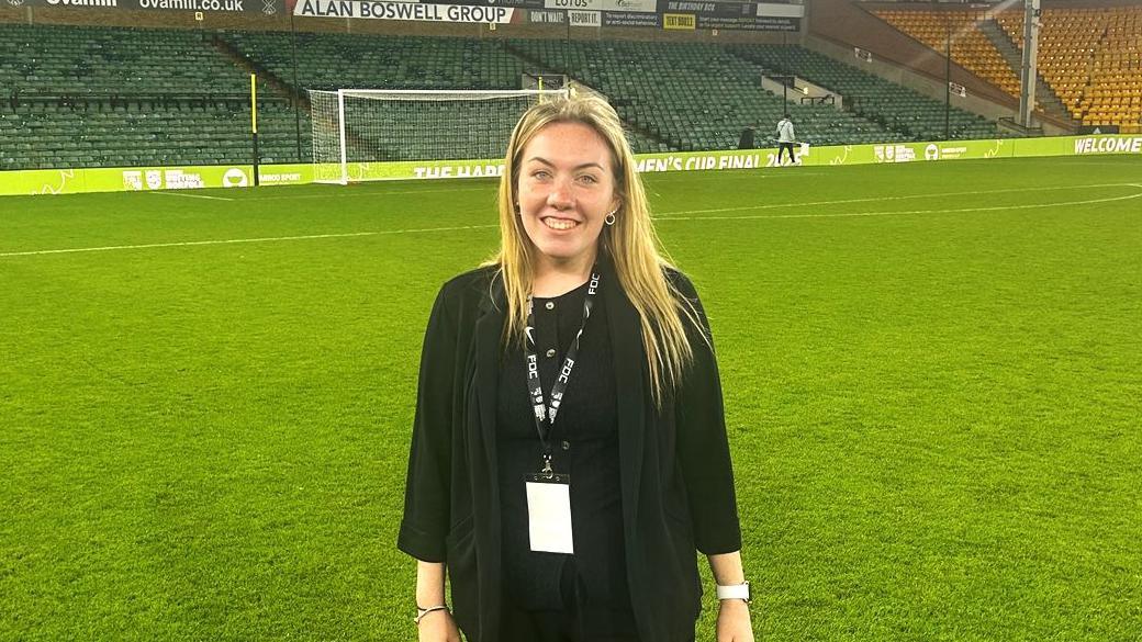 Chloe Moulton is standing on the pitch at Norwich City's Carrow Road ground. Behind her is a a goal and a stand. She's wearing a black cardigan over a black top.