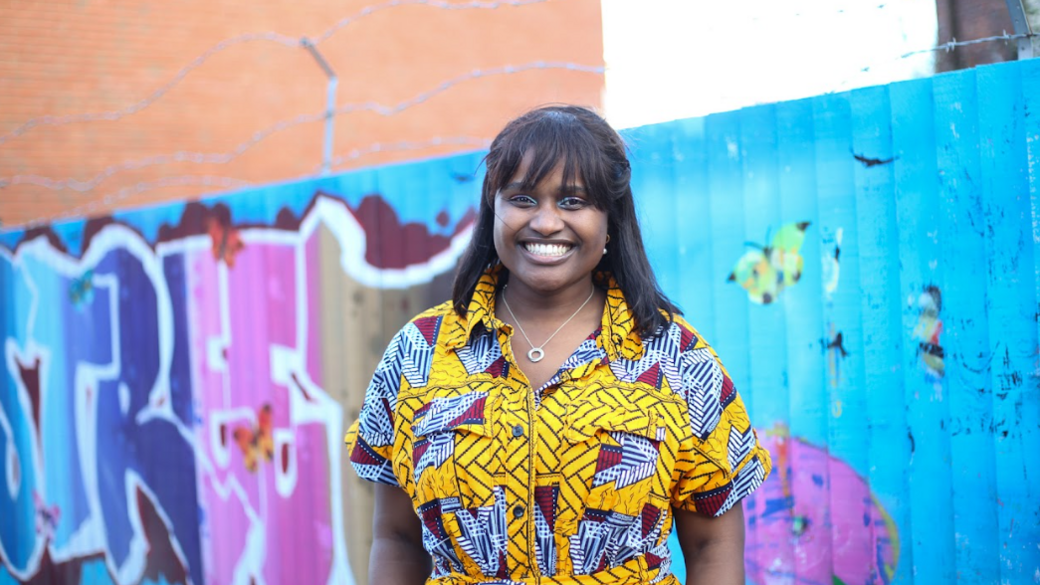 Ms Ndibwirende in a brightly-coloured yellow dress standing, smiling, in front of a fence which has street art painted on it.