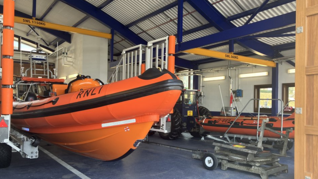 Two orange lifeboats inside a building which has blue rafters. One boat is larger than the other. There is also a blue tractor next to the smaller boat