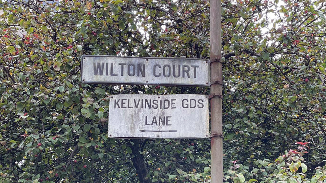 Weathered grey sign saying Wilton Court, along with a sign saying Kelvinside Gds Lane. There are trees behind it.