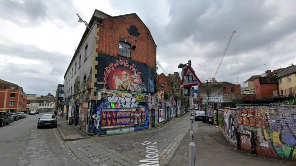 A red brick building is covered in graffiti on the corner of two side streets. A two-way sign is in view and cars parked.