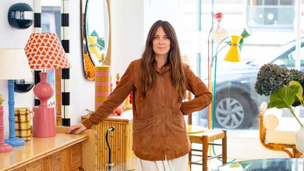 A photo of Natalie Tillison - a woman with long brown hair standing in a brightly lit interior filled with colourful furniture and decor. She wears a brown suede jacket and a white skirt and is leaning on a wooden sideboard. The design studio features vibrant lamps, striped columns, mirrors and rattan furniture, with large windows showing a city street, with a black car parked outside.