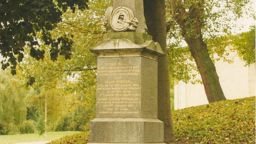 A stone memorial with gold lettering in a green wooded area of a cemetery.