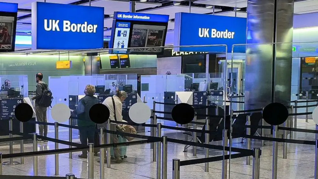 An airport border control area with large information screens overhead and booths for passport officials. There are lanes cordoned off for queuing, but only three people waiting to pass through.