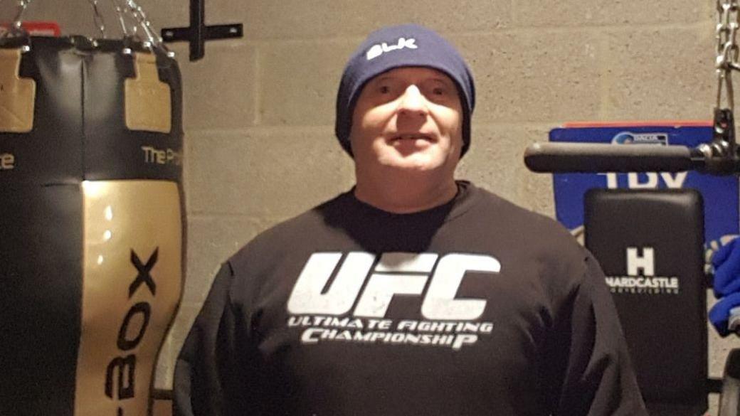 Steve Scott in his home gym with the punch bag behind him and dressed in a hoodie that reads 'UFC'