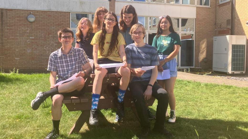 A group of students - three boys and four girls - sitting on a picnic bench outside a school building, holding their results.
