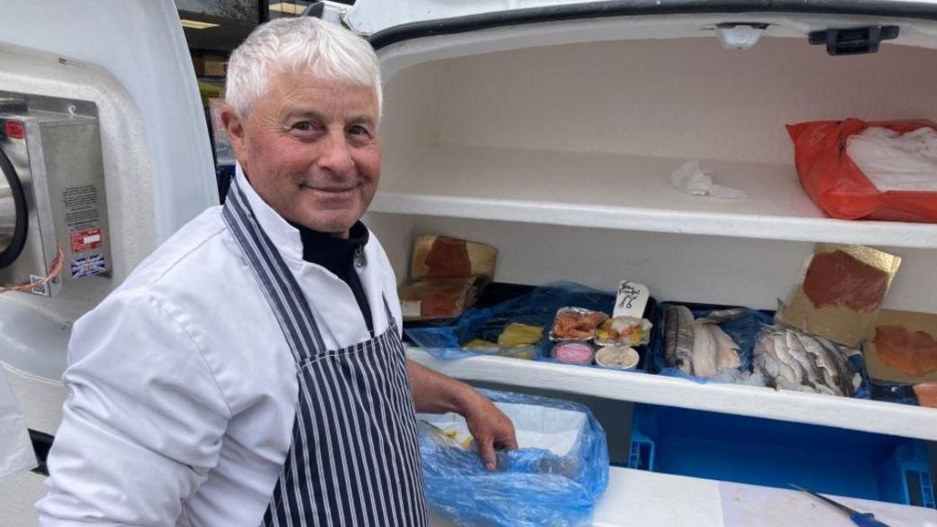 Fishmonger Graham Garrod , wearing a white jacket under a striped apron, is standing in front of a counter containing an array of seafood products.