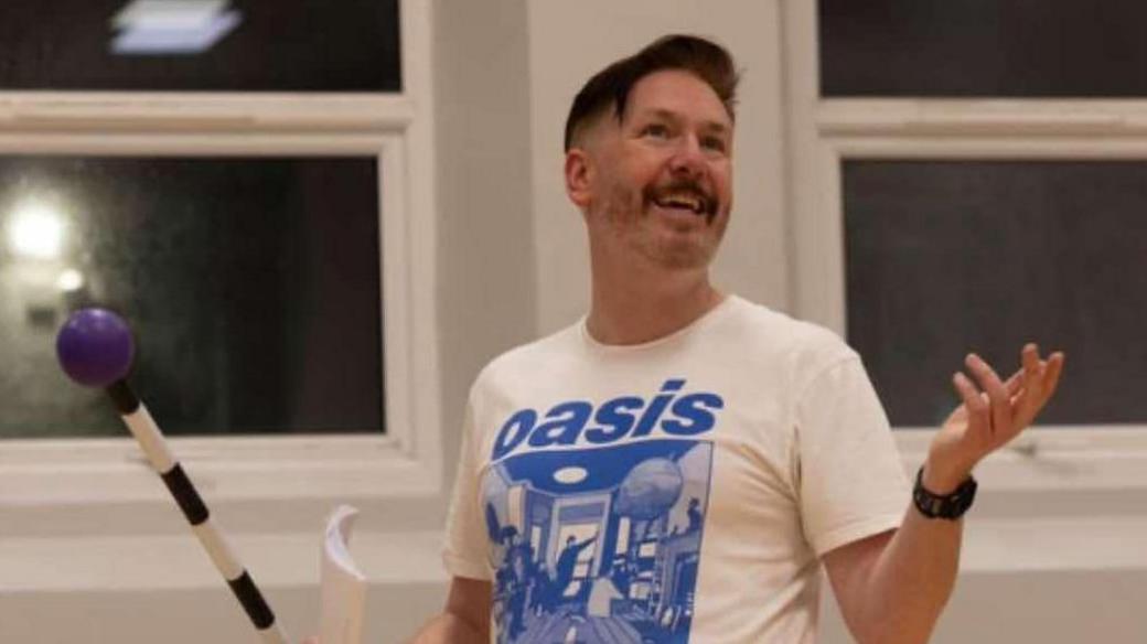  Alan McClarty is seen in an Oasis shirt during a rehearsal for a performance. He has a large cane in his hand.